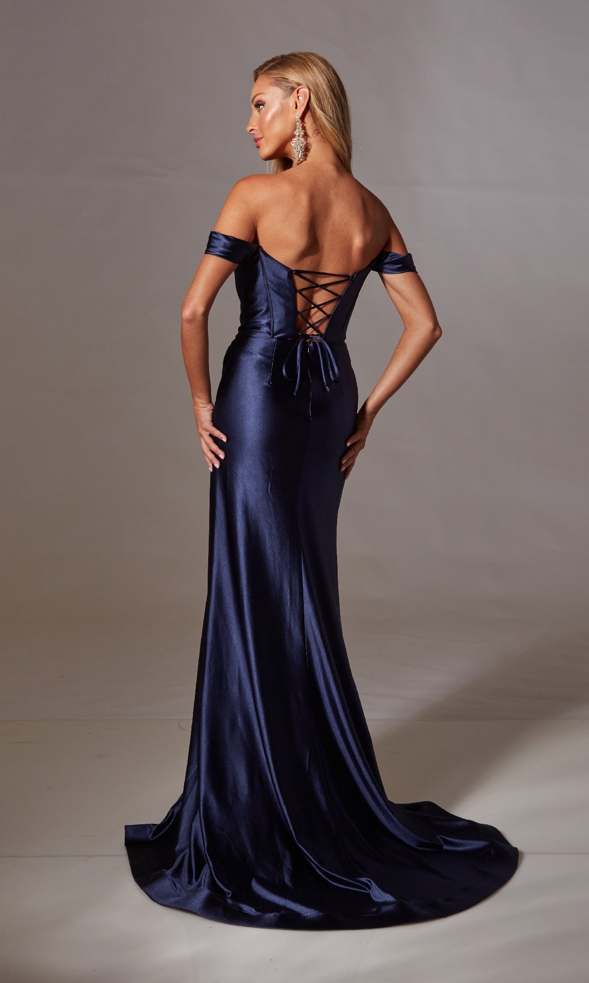 A woman faces away, wearing the Aerie Couture 240 Lace-Up Long Corset Prom Dress featuring an off-the-shoulder design, dark blue satin fabric, a corset-style bodice, and a long trailing skirt.
